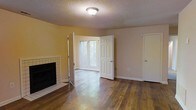 This single-story residence offers a well-organized and comfortable layout spread across multiple interconnected rooms. Rich, warm-toned hardwood flooring runs throughout the main living areas, lending a cohesive and inviting character to the space. The living room serves as a central gathering area, highlighted by a classic tile-surround fireplace that anchors the room with both warmth and style. Windows allow natural light to filter in, brightening the warm yellow-toned walls and textured ceilings. The kitchen is thoughtfully positioned and features ample counter space and cabinetry, providing a functional and organized cooking environment. The floor plan, as seen from above, reveals a spacious and somewhat L-shaped configuration, with clearly defined zones for living, dining, and private areas. Hallways connect the various rooms, which include spaces that can serve as bedrooms or flexible-use rooms, each benefiting from the consistent hardwood flooring and neutral wall tones. The overall ambiance is warm and homey, with a traditional aesthetic reinforced by paneled interior doors, textured ceilings, and classic finishes throughout. The layout prioritizes both functionality and comfort, making it a practical and welcoming living space.