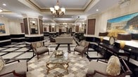 This sophisticated single-floor space exudes timeless elegance through its meticulously crafted classical interior design. The centerpiece of the space is a grand, open lounge area anchored by a striking black-and-white polished marble floor, which transitions seamlessly into a detailed geometric patterned area rug in warm, neutral tones. Overhead, ornate brass chandeliers with frosted glass shades cast a warm, inviting glow across the room, complemented by recessed lighting embedded within coffered tray ceilings adorned with intricate crown molding and decorative gold-toned trim. The seating arrangement features plush, taupe upholstered sofas and armchairs with dark wood frames and embroidered accent cushions, centered around a sculptural gold-framed glass coffee table. Dark wood wall panels, framed artwork, arched architectural detailing, and decorative wall sconces contribute to the space's refined, stately atmosphere. A console table with a statement lamp and decorative accessories lines one wall, adding layered depth to the room's composition. The generous proportions of the space, along with the tall ceilings and open layout, create an expansive and welcoming ambiance suited for formal reception or upscale lounge use.