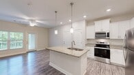 Located at 12102 Reserve Manor Cir in Chester, VA, this single-floor, one-bedroom, one-bathroom residence offers a thoughtfully designed open-concept layout that maximizes its comfortable footprint. The combined kitchen, dining, and living area serves as the heart of the home, highlighted by a central kitchen island with a granite countertop and built-in sink, illuminated by two chrome pendant lights. White shaker-style cabinetry pairs seamlessly with granite countertops, while a full suite of stainless steel appliances—including a refrigerator, range, and over-the-range microwave—adds a modern, polished touch. Wood-look flooring flows throughout the main living areas, complementing the neutral, light-toned walls. The living room benefits from a ceiling fan and a glass door that opens to an outdoor area, inviting natural light. A hallway connects the main living space to the private bedroom, which features plush carpet, a ceiling fan, and a window with views of the surrounding greenery. The bathroom includes a granite-topped vanity with an undermount sink, a tub-shower combination, and matching wood-look flooring. A dedicated closet and hallway round out this efficiently organized and move-in-ready residence.