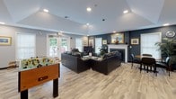 This single-floor community space is thoughtfully designed to offer a welcoming and versatile environment for socializing and recreation. The main lounge area is anchored by a classic white-mantled fireplace set against a bold navy accent wall, adorned with gold-framed artwork and decorative mirrors. Plush black leather sofas and armchairs are arranged to encourage conversation and relaxation, all resting on light-toned hardwood-style flooring that runs throughout the space. Recessed lighting and a vaulted ceiling section lend an open, airy feel to the room, while large windows and French doors flood the interior with natural light and provide direct access to an outdoor deck furnished with seating for al fresco enjoyment.

A dedicated game room adjacent to the lounge features a full-size pool table and a foosball table, offering ample entertainment options. A compact kitchenette is also part of the layout, equipped with light wood cabinetry, a sink, a full-size refrigerator, and a coffee maker — ideal for serving refreshments. A small meeting or work area with chairs rounds out the functional offerings of this space. Together, these areas create a multifunctional community hub that balances comfort, recreation, and convenience in a clean, modern setting.