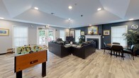 This single-floor community space is thoughtfully designed to offer a welcoming and versatile environment for socializing and recreation. The main lounge area is anchored by a classic white-mantled fireplace set against a bold navy accent wall, adorned with gold-framed artwork and decorative mirrors. Plush black leather sofas and armchairs are arranged to encourage conversation and relaxation, all resting on light-toned hardwood-style flooring that runs throughout the space. Recessed lighting and a vaulted ceiling section lend an open, airy feel to the room, while large windows and French doors flood the interior with natural light and provide direct access to an outdoor deck furnished with seating for al fresco enjoyment.

A dedicated game room adjacent to the lounge features a full-size pool table and a foosball table, offering ample entertainment options. A compact kitchenette is also part of the layout, equipped with light wood cabinetry, a sink, a full-size refrigerator, and a coffee maker — ideal for serving refreshments. A small meeting or work area with chairs rounds out the functional offerings of this space. Together, these areas create a multifunctional community hub that balances comfort, recreation, and convenience in a clean, modern setting.