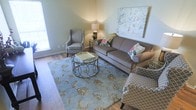 This single-floor space showcases a refined and inviting interior design centered around comfort and classic style. The living area serves as the heart of the space, featuring rich hardwood flooring that adds warmth and a sense of timeless elegance throughout. The room is anchored by a plush, taupe upholstered sofa with nailhead trim detailing, complemented by a pair of coordinating patterned armchairs in a soft lattice print that add visual interest without overwhelming the palette. A large, intricately patterned area rug in muted blue and gold tones grounds the seating arrangement, adding texture and a gentle pop of color to the neutral setting. A decorative wrought-iron coffee table with a tray top serves as a stylish and functional centerpiece. Soft, ambient lighting from a table lamp placed on an ornate side table creates a cozy and welcoming atmosphere. The walls are painted in a light, soothing tone that enhances the airy feel of the room, while a large, vibrant botanical artwork displayed prominently above the sofa draws the eye and adds an artistic focal point. Natural light filters in through a window, further brightening the space and contributing to its warm, comfortable ambiance.