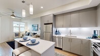 This thoughtfully designed two-bedroom, one-bathroom residence at 59 S L St in Livermore, California, offers a contemporary living experience within a compact, efficiently utilized single-floor layout. The open-concept kitchen serves as the centerpiece of the unit, showcasing grey flat-front cabinetry extending to the ceiling, white quartz countertops, a classic subway tile backsplash with under-cabinet lighting, and a full suite of stainless steel appliances including a gas range, built-in microwave, dishwasher, and refrigerator. A central kitchen island with seating provides additional prep space and casual dining. Light wood-style flooring flows throughout the main living areas, creating a warm, cohesive aesthetic. The living room benefits from natural light through windows overlooking an outdoor patio, accessible via a glass-paneled door. The primary bedroom features a large window with patio views and soft carpeted flooring. A hallway connects both bedrooms to a shared closet. The full bathroom offers a tiled tub-shower combination, a sleek vanity with grey cabinetry, and large-format floor tiles. The overall ambiance is clean, modern, and bright.