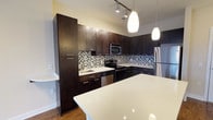 This single-floor space showcases a well-appointed kitchen designed with a contemporary aesthetic. Dark espresso-toned cabinetry extends from floor to ceiling, providing generous storage while creating a bold visual contrast against the light quartz countertops. The counters offer ample workspace and continue onto a large central island, which serves as a functional focal point ideal for meal preparation or casual dining.

A mosaic tile backsplash in neutral tones of gray, white, and black runs the length of the kitchen, adding texture and visual interest between the upper and lower cabinets. Stainless steel appliances, including a range, microwave, dishwasher, and refrigerator, complement the modern design while ensuring full functionality.

Overhead, a combination of recessed track lighting and elegant teardrop-shaped pendant lights illuminate the space effectively. The hardwood flooring grounds the kitchen warmly, balancing the cooler tones of the cabinetry and stone surfaces. Overall, the kitchen presents a cohesive, upscale design that blends practicality with refined style.