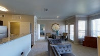 This single-story space in Grapevine, Texas, presents a well-appointed interior designed with a cohesive neutral color palette of warm grays and soft whites. The living area features plush carpeting underfoot and is furnished with tufted velvet seating that adds a touch of sophistication to the space. Crown molding runs along the ceiling line, lending a refined architectural detail throughout the open layout.

The dining area is positioned toward the rear of the space and centers around a glass-top table with upholstered accent chairs. A decorative quatrefoil mirror and an ornate floral arrangement serve as a focal point on the wall, adding visual interest. Natural light enters through a window fitted with blinds nearby, brightening the area without sacrificing privacy.

Recessed lighting in the ceiling provides consistent, even illumination throughout. A partial wall with a granite countertop edge hints at an adjacent kitchen space, suggesting a practical open-concept flow. A built-in shelving unit is visible down the hallway, adding storage without compromising the clean aesthetic. The overall ambiance is polished, comfortable, and move-in ready.