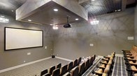 This elegant private screening room delivers an immersive cinematic experience within a thoughtfully designed single-level space. The room is anchored by a large, wall-mounted projection screen at the front, paired with a ceiling-mounted projector that ensures a high-quality viewing experience from every seat. Multiple rows of plush, theater-style seating are arranged in a tiered, stadium configuration, upholstered in rich brown and tan tones with armrests featuring integrated cup holders — combining comfort with practicality. The ceiling is a standout architectural feature, showcasing a combination of a decorative patterned tile section and a smooth, coffered central recess fitted with recessed can lighting. Two ornate crystal chandeliers flank the projector, adding a touch of refined glamour and warm ambient illumination to the space. The walls are finished in a warm, neutral gray tone accented by subtle artistic detailing, contributing to the room's upscale yet inviting atmosphere. Striped carpeting runs along the floor, reinforcing the tiered layout while adding visual texture. Overall, the space strikes a balance between the functionality of a professional screening environment and the aesthetic warmth of a luxury entertainment venue.