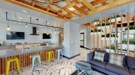 This thoughtfully designed single-floor communal space blends industrial, natural, and contemporary elements into a cohesive and inviting environment. The standout flooring features large-format white tiles with dramatic black veining, creating a bold visual foundation throughout the space.

The expansive kitchen is a focal point, outfitted with stainless steel appliances including a built-in cooktop, range hood, microwave, and refrigerator. Light wood cabinetry pairs with gray quartz countertops, while the walls are adorned with intricate embossed white decorative tiles that extend to the ceiling. Ornate matching ceiling tiles and Edison bulb pendant lighting suspended on industrial track fixtures complete the kitchen's eclectic aesthetic.

Adjacent to the kitchen, a bar-height counter with colorful metal stools provides casual seating, making it ideal for social gatherings. A wall-mounted television adds entertainment functionality to this area.

The lounge zone features a plush gray sofa, patterned throw pillows, and a round tufted ottoman, all anchored by a soft area rug. A slatted wood room divider with integrated planters provides a natural, green partition between areas, reinforcing the biophilic design theme.

Overhead, exposed natural wood beam ceilings with a coffered grid pattern add warmth and architectural interest, while an industrial-style cage ceiling fan circulates air throughout. Large windows allow natural light to fill the space, enhancing its welcoming, modern ambiance.