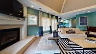 Located at 2502 Manor Dr in Fredericksburg, VA, this single-story space presents a cohesive, design-forward interior defined by a striking teal and navy color palette carried consistently throughout. The open floor plan connects the main living, dining, and kitchen areas, creating a fluid and functional layout ideal for both everyday use and entertaining.

The living area features soaring vaulted ceilings with a coffered detail, recessed lighting, and large windows dressed with layered Roman shades and floor-length curtains that invite abundant natural light. A white fireplace surround anchors one wall, while light hardwood flooring runs throughout the space, adding warmth and continuity.

The dining area is distinguished by a tray ceiling, an oversized pendant light, and a large round table surrounded by upholstered chairs. Natural greenery and wall art enhance the refined atmosphere. An entryway adorned with a geometric lantern pendant provides a welcoming transition into the open interior.

The kitchen is well-appointed with dark espresso cabinetry, white countertops, stainless steel appliances, and a peninsula with bar-height seating. A window above the sink provides garden views, and pendant lighting above the peninsula adds a polished finishing touch.