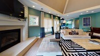 Located at 2502 Manor Dr in Fredericksburg, VA, this single-story space presents a cohesive, design-forward interior defined by a striking teal and navy color palette carried consistently throughout. The open floor plan connects the main living, dining, and kitchen areas, creating a fluid and functional layout ideal for both everyday use and entertaining.

The living area features soaring vaulted ceilings with a coffered detail, recessed lighting, and large windows dressed with layered Roman shades and floor-length curtains that invite abundant natural light. A white fireplace surround anchors one wall, while light hardwood flooring runs throughout the space, adding warmth and continuity.

The dining area is distinguished by a tray ceiling, an oversized pendant light, and a large round table surrounded by upholstered chairs. Natural greenery and wall art enhance the refined atmosphere. An entryway adorned with a geometric lantern pendant provides a welcoming transition into the open interior.

The kitchen is well-appointed with dark espresso cabinetry, white countertops, stainless steel appliances, and a peninsula with bar-height seating. A window above the sink provides garden views, and pendant lighting above the peninsula adds a polished finishing touch.