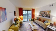 This single-floor space showcases a thoughtfully designed living area that balances comfort and personality. The living room serves as the centerpiece, immediately drawing attention with its bold color palette — striking orange curtains frame large sliding glass doors that flood the room with natural light and offer a view of the greenery outside. The sliding doors also provide direct access to a private balcony, extending the sense of openness beyond the interior.

The room features a plush carpeted floor accented by a vibrant floral-patterned area rug, adding warmth and visual interest underfoot. A cream-toned sofa anchors the seating area, complemented by a mustard-yellow armchair and a matching yellow console that introduces a cheerful, retro-inspired aesthetic. A clean white coffee table sits at the center, providing a functional yet minimalist contrast to the lively surrounding décor.

Wall art, decorative throw pillows in coordinating earth tones, and carefully placed accent lamps contribute to the cozy and curated ambiance. The textured ceiling adds a classic architectural detail overhead. Overall, this space exudes a warm, welcoming character with a distinctive design sensibility that blends bold color choices with comfortable, everyday living.