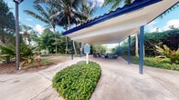Located at 6116 Yellow Sun Drive in Lantana, Florida, this outdoor covered pavilion offers a welcoming communal gathering space nestled within a beautifully landscaped tropical setting. The structure is defined by its distinctive blue-trimmed, corrugated roof supported by bold blue steel columns, providing reliable shade over a smooth concrete floor. Picnic tables situated beneath the canopy make this an ideal spot for outdoor dining, relaxation, or community events. A centrally placed informational signpost adds a practical, community-oriented touch to the space. The pavilion is framed by lush, mature palm trees and dense tropical greenery, creating a vibrant natural backdrop that enhances the Florida ambiance. Manicured low shrubs border the approach to the structure, adding a polished, well-maintained appearance. Glimpses of a nearby water feature or lake are visible through the surrounding foliage, further enriching the scenic outdoor environment. The open-air design allows for natural breezes and abundant natural light to flow freely through the space, making it a comfortable retreat throughout the day. This pavilion serves as a charming and functional outdoor amenity within its South Florida community.