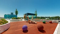 This rooftop amenity space in Surrey, BC offers a thoughtfully designed outdoor retreat high above the city, combining recreational, social, and green spaces under open skies. The layout is divided into distinct zones, each serving a specific purpose while contributing to the overall cohesive design.

A standout feature is the children's play area, surfaced with a resilient red rubberized flooring for safety and comfort. The area is outfitted with natural wood sculptural play structures and oversized colorful spheres, creating an engaging and imaginative environment for younger residents. Curved concrete seating walls with integrated lighting border the play zone, providing both structure and casual seating for observers.

Adjacent to the play area, a paved terrace features a BBQ station complete with wooden prep stations and a fire pit, ideal for outdoor gatherings and entertaining. Large cylindrical concrete planters add greenery and visual interest throughout this zone, while glass-paneled enclosures provide shelter from the elements.

Lush raised garden beds and young trees are strategically planted across the rooftop, softening the urban environment and enhancing the sense of nature. Glass railings along the perimeter ensure unobstructed, panoramic views of Surrey's dynamic skyline, including neighboring high-rise towers and the surrounding treeline, making this rooftop a versatile and vibrant community amenity.