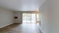 Located at 38730 Lexington St in Fremont, California, this single-floor residence offers a practical and comfortable layout with a clean, modern aesthetic throughout. The living area is a standout feature, showcasing warm wood-look hardwood flooring, neutral gray walls, and a large sliding glass door fitted with vertical blinds that floods the space with natural light and provides direct access to an outdoor patio area with views of surrounding greenery. A wall-mounted AC unit adds functional convenience to the living space. The open transition between the living and dining areas is defined by a mix of wood-look and tile flooring, with a pass-through window detail that connects to adjacent spaces and enhances the open feel of the layout. The galley-style kitchen is well-appointed with white cabinetry, granite-look countertops, a stainless steel refrigerator, dishwasher, and range, all set atop light ceramic tile flooring. The bedroom features the same rich wood-look flooring, a ceiling fan with light fixture, and a window offering natural light and outdoor views. The bathroom hallway includes matching countertops and tile flooring consistent with the rest of the home. Overall, the space presents a cohesive, move-in-ready interior with thoughtful finishes throughout.