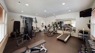 Located at 6614 North Cedar Lane in Spokane, Washington, this dedicated fitness center is a well-appointed, single-floor space designed to support a comprehensive workout experience. The room is finished with clean white walls and ceilings accented by recessed lighting, creating a bright and welcoming environment. Durable rubber flooring covers the entire workout area, providing both safety and functionality.

The space is generously equipped for strength training, featuring a multi-station cable machine, a Matrix adjustable weight bench, a full dumbbell rack ranging across multiple weight increments, and a selection of kettlebells and medicine balls neatly organized along the walls. A wall-mounted flat-screen television adds an entertainment element to the space, along with a wall clock and a coat hook station near the entrance.

For cardiovascular training, the facility includes multiple treadmills, an elliptical machine, and a stationary bike, all positioned to take advantage of the natural light streaming through the numerous windows fitted with warm wood-toned blinds. A bulletin board and informational materials are also available for users. The layout is thoughtfully organized to allow ample open floor space between equipment, ensuring comfort and ease of movement throughout the facility.