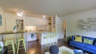 This single-floor space showcases an open-concept layout that seamlessly connects the kitchen and living area, creating a cohesive and functional living environment. The kitchen is outfitted with white cabinetry, white appliances including a wall oven, microwave, and refrigerator, and is well-lit by under-cabinet lighting. A peninsula-style breakfast bar extends from the kitchen, providing casual seating for two or three bar stools, which are finished in a vibrant lime green — a detail echoed throughout the space's décor. Rich, dark hardwood-style flooring runs throughout, grounding the bright white walls and cabinetry with a warm contrast. The living area flows naturally from the kitchen, featuring a ceiling fan, a textured ceiling, and comfortable seating in bold blue tones. A striped area rug in green and white adds a playful touch. A doorway visible from the main area leads to additional rooms, suggesting a practical and well-organized floor plan. The overall ambiance is light, airy, and cheerfully styled, with a cohesive palette of white, green, and blue lending the space a fresh, coastal-inspired character.