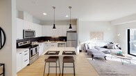 This well-appointed single-floor residence offers a thoughtfully designed open-concept layout that seamlessly connects the kitchen, dining, and living areas. The kitchen is a standout feature, showcasing sleek white cabinetry extending to the ceiling, a dark tile backsplash, stainless steel appliances including a full-size refrigerator, electric range, built-in microwave, and dishwasher, along with a central island with bar seating illuminated by pendant lighting. Light hardwood-style flooring runs throughout the main living areas, enhancing the bright and contemporary feel.

The living space flows naturally from the kitchen, offering a comfortable and open atmosphere. Two well-sized bedrooms are finished with plush carpeting and feature large windows that invite natural light. One bedroom includes direct access to a full bathroom with a tub/shower combination, a wide vanity with ample counter space, and a walk-in closet visible through the doorway. The second bedroom also enjoys generous proportions and large windows.

A dedicated laundry area houses a stacked Whirlpool front-loading washer and dryer alongside wire shelving for additional storage. The unit's neutral color palette, recessed lighting, and clean architectural lines contribute to a cohesive, modern aesthetic throughout.