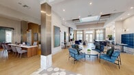 Located at 2730 Welaunee Blvd in Tallahassee, FL, this sophisticated single-floor community clubhouse and leasing space offers a generously sized, open-concept layout designed to impress. The interior greets visitors with a striking reception area featuring a sleek wood-toned desk, bold geometric patterned tile flooring, and metallic accent columns with marble bases that anchor the space elegantly.

The open living area showcases soaring tray ceilings with dark wood beam detailing, recessed lighting, and large French doors that flood the space with natural light while offering outdoor views. A floor-to-ceiling tile fireplace paired with a mounted TV serves as a dramatic focal point, flanked by plush navy blue and neutral-toned seating arrangements. An adjacent bar-height island with a white marble countertop and dark cabinetry provides a stylish gathering spot.

The dining area features a bold geometric-patterned accent wall, pendant globe lighting, and a mix of intimate seating configurations. A cozy lounge room offers a warm, layered aesthetic with a built-in wood-slat shelving unit, coffered ceiling details, and a large sectional sofa. Throughout the space, light hardwood-style flooring, curated artwork, and carefully selected furnishings in navy, gray, and earth tones create a cohesive, modern, and welcoming ambiance.