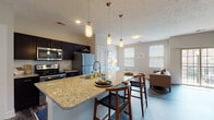 This single-floor residence is designed with a contemporary open-concept layout that seamlessly connects the kitchen, dining, and living areas. The kitchen is a standout feature, centered around a generous granite-topped island with bar seating and illuminated by a trio of stylish pendant lights. Dark espresso cabinetry contrasts elegantly with the light granite countertops, while stainless steel appliances—including a refrigerator, range, and microwave—provide a sleek, modern finish. A white subway tile backsplash adds a clean, classic touch.

The open living area flows naturally from the kitchen, featuring dark hardwood-style flooring and vaulted ceilings in select areas that enhance the sense of space. Multiple windows fitted with horizontal blinds allow generous natural light throughout, while sliding glass doors in the living room open to a private balcony, offering outdoor views.

The bedroom is carpeted and well-lit by two large windows, creating a calm and comfortable retreat. The bathroom is finished with light tile flooring, a single-sink vanity with neutral cabinetry, a soaking tub with shower, and convenient access to an open walk-in closet with built-in shelving. The overall ambiance is clean, bright, and modern throughout.