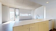 Located at 11461 Euclid Ave in Cleveland's University Circle neighborhood, this single-floor unit showcases a clean, modern aesthetic defined by exposed concrete ceilings and an open, airy layout. The living and dining areas are anchored by dark wood-look flooring and generous floor-to-ceiling windows, allowing natural light to pour in and offering views of the surrounding urban streetscape. Subtle cove lighting along the ceiling adds a warm ambiance to the open common areas.

The kitchen is thoughtfully designed with warm-toned cabinetry, white solid-surface countertops, and a kitchen island with an integrated sink. A galley-style cooking area provides full appliance accommodation, including a range, microwave, and dishwasher, with ample cabinetry for storage. Sliding glass-panel doors provide access to utility connections nearby.

The bedroom features plush carpeting and large windows that invite in plenty of daylight, framing views of the neighborhood beyond. The bathroom is finished in crisp white tile, complete with a glass-enclosed walk-in shower, modern fixtures, and a clean vanity with integrated sink — delivering a minimalist, spa-like feel. Throughout the unit, the consistent use of neutral tones and quality materials creates a cohesive, contemporary living environment.