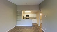 This single-floor space features an open-concept design that seamlessly connects the living area to the kitchen. The living space is finished with warm hardwood-style flooring that extends throughout, complemented by neutral taupe-toned walls and white baseboards that create a clean, cohesive aesthetic. A standout architectural detail is the built-in shelving unit integrated into the half-wall partition, offering both display and storage functionality while defining the boundary between the living and kitchen areas.

The kitchen is equipped with white shaker-style cabinetry, dark countertops, and a full suite of black appliances, including a range, microwave, and refrigerator. Overhead lighting illuminates the kitchen workspace effectively. The peninsula-style layout provides additional counter space and casual seating potential.

A doorway visible from the main living area leads to additional rooms, suggesting further private living spaces beyond the open common areas. The overall ambiance is bright and functional, with a modern yet approachable design sensibility suited for everyday living.