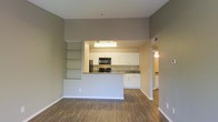 This single-floor space features an open-concept design that seamlessly connects the living area to the kitchen. The living space is finished with warm hardwood-style flooring that extends throughout, complemented by neutral taupe-toned walls and white baseboards that create a clean, cohesive aesthetic. A standout architectural detail is the built-in shelving unit integrated into the half-wall partition, offering both display and storage functionality while defining the boundary between the living and kitchen areas.

The kitchen is equipped with white shaker-style cabinetry, dark countertops, and a full suite of black appliances, including a range, microwave, and refrigerator. Overhead lighting illuminates the kitchen workspace effectively. The peninsula-style layout provides additional counter space and casual seating potential.

A doorway visible from the main living area leads to additional rooms, suggesting further private living spaces beyond the open common areas. The overall ambiance is bright and functional, with a modern yet approachable design sensibility suited for everyday living.
