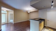This single-floor loft-style unit showcases a compelling blend of industrial and contemporary design. Exposed concrete ceilings with visible steel beams and large metallic ductwork run throughout the space, lending it a distinct urban character. Rich, dark hardwood flooring flows seamlessly across the main living areas, providing a warm contrast to the industrial overhead elements.

The open-concept kitchen features light maple cabinetry, dark granite countertops, and black appliances including a refrigerator, gas range, and built-in microwave. Track lighting illuminates the kitchen efficiently, while tile flooring defines the cooking zone. A peninsula counter creates a natural divide between the kitchen and living area.

The spacious living area is bathed in natural light from multiple large windows and a glass-panel door that opens to an outdoor space, offering views of the surrounding urban environment. Built-in shelving adds both function and visual interest to one of the living room walls.

A hallway with pendant lighting connects the main living space to the private areas, where a full bathroom features a combined tub and shower, a vanity with maple cabinetry, and hardwood flooring. Additional closet storage is also accessible from the hallway. The overall ambiance is open, bright, and distinctly loft-inspired.