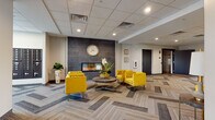 This thoughtfully designed single-floor space offers a comprehensive suite of amenities within a modern, cohesive interior. The welcoming lobby and lounge area immediately sets the tone, showcasing bold yellow tufted armchairs and a sofa arranged around a sleek coffee table atop herringbone-patterned carpet tiles that run throughout the space. A decorative accent wall featuring a geometric tile pattern serves as the focal point, framing a linear electric fireplace beneath a large wall clock, while curated artwork adds character to the neutral walls. Natural light filters in through multiple windows, complementing the recessed ceiling lighting overhead. Adjacent to the lounge, a well-equipped kitchenette features dark wood cabinetry, granite countertops, a large center island with pendant lighting, and stainless steel appliances including a full-sized refrigerator. A separate dining nook accommodates a round table with seating for four. The fitness room offers a varied selection of cardio machines, a multi-function cable system, free weights, and medicine balls, with wall mirrors enhancing the sense of space. An accessible restroom with light tile finishes and grab bars, along with elevator access and stairwell entry, round out the functional offerings of this well-planned space.