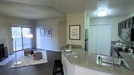 This single-floor residence offers a thoughtfully designed open-concept layout that connects the kitchen and living areas into one cohesive, functional space. The kitchen is a standout feature, showcasing a generous peninsula with speckled granite-style countertops and a breakfast bar with seating, making it ideal for casual dining or entertaining. Stainless steel appliances, including a full-size refrigerator, add a modern, polished touch to the cooking space. The countertops extend to create ample prep and serving areas, complemented by clean white cabinetry that brightens the room. Rich, dark hardwood-style flooring spans the main living areas, providing a warm contrast to the neutral gray walls throughout. The open living room offers generous space for furniture arrangement, with a cozy area rug and decorative accents adding personality to the room. Framed artwork on the walls enhances the inviting atmosphere, while a large window allows natural light to fill the space. The textured ceiling adds a subtle architectural detail throughout. Overall, the space presents a clean, contemporary aesthetic with a practical, open layout well-suited for comfortable everyday living.