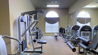 This single-floor fitness space is designed to support a comprehensive strength training regimen. The room features warm, neutral-toned walls and carpeted flooring, creating a functional yet comfortable atmosphere. A standout architectural detail is the arched window adorned with a decorative valance, which allows natural light to filter into the space and adds a refined visual element to the room. Overhead, a recessed fluorescent light fixture ensures consistent illumination throughout. The space is outfitted with multiple cable and resistance machines positioned along the perimeter, catering to a range of upper and lower body exercises. A dumbbell rack with a variety of free weights provides additional versatility for strength training routines. A large wall-mounted mirror along one side of the room enhances the sense of space and allows users to monitor their form during workouts. The layout is thoughtfully arranged to allow for ease of movement between equipment. Overall, the space balances practical fitness functionality with an inviting, well-maintained ambiance suited for focused, effective workouts.
