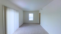 This single-story space presents a clean, minimalist interior centered around a spacious open room. The room features neutral beige carpeting that extends throughout, complemented by smooth, light-colored walls that contribute to an airy and understated aesthetic. A centrally positioned window allows natural light to filter in, brightening the interior and enhancing the sense of openness. Vertical blinds on the left side suggest an additional window or sliding door, offering further potential for light and access. The ceiling is standard height and smooth-finished, maintaining the room's simple, uncluttered character. A doorway visible on the right side of the room indicates a connected adjoining space, suggesting a functional layout that extends beyond this primary room. The overall ambiance is neutral and move-in ready, offering a blank canvas well-suited for a variety of furnishing arrangements and interior design preferences. The combination of soft flooring, light walls, and natural illumination makes this a versatile and welcoming space.