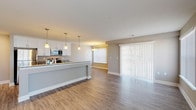 This single-floor unit offers a thoughtfully designed open-concept layout that seamlessly connects the kitchen and living areas. The kitchen is a standout feature, showcasing crisp white shaker cabinetry, granite countertops, and a generously sized peninsula island ideal for casual dining or meal prep. Pendant lighting adds warmth above the island, while a full suite of stainless steel appliances — including a gas range, built-in microwave, and refrigerator — completes the space. Wood-look plank flooring runs throughout the main living area, contributing to a clean and cohesive aesthetic. Large sliding glass doors and additional windows dressed with vertical blinds allow natural light to fill the open living space. The bedroom features soft wall-to-wall carpeting and a bright window, providing a comfortable and quiet retreat. The bathroom is elegantly finished with large-format gray stone-look tile surrounding a soaking tub, complete with a built-in niche for added convenience. A dedicated laundry area houses a stacked washer and dryer unit with wire shelving for storage. Neutral gray tones and white trim throughout the space create a modern, move-in-ready atmosphere.