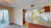 This single-floor residence offers a practical and comfortable layout, with distinct living, kitchen, and bathroom areas connected by an open, flowing floor plan. The kitchen is a functional centerpiece, featuring rich medium-toned wood cabinetry both above and below the counters, granite-patterned countertops, a stainless steel sink, and white appliances including a coil electric range with overhead ventilation, a dishwasher, and a refrigerator. A peninsula counter extends into the adjacent area, creating a natural transition between the kitchen and living spaces. The living area is carpeted and bright, illuminated by a ceiling fan with integrated lighting and a large sliding glass door fitted with vertical blinds that opens to an outdoor patio, offering views of greenery beyond. The entryway features wood-style flooring, providing a clean threshold from the exterior door into the carpeted main living space. A side room, visible through a connecting passage, provides additional flexible living space with its own carpeting and natural light. The bathroom is neatly finished with warm wood cabinetry, a granite-patterned countertop, an undermount sink, a large framed mirror with overhead vanity lighting, and wood-style flooring. Textured ceilings and neutral warm-toned walls carry throughout the space, lending a consistent and cohesive aesthetic.