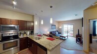 This single-floor residence offers a thoughtfully designed open-concept layout that seamlessly connects the kitchen, dining, and living areas. The kitchen is a standout feature, showcasing rich dark-wood cabinetry, granite countertops, and a full suite of stainless steel appliances including a refrigerator, range, and microwave. A peninsula with bar seating serves as both a functional workspace and a social hub, illuminated by a trio of elegant pendant lights. Hardwood flooring runs throughout the kitchen and entry areas, transitioning to plush carpet in the living room and bedrooms. The living area is bright and welcoming, with natural light streaming in through a sliding glass door that opens to a private balcony. The two bedrooms are comfortably sized, each featuring windows for natural light and sliding closet doors with built-in storage. The full bathroom is clean and functional, featuring a vanity with dark cabinetry, a combined tub and shower, and light wood-tone flooring. Track lighting in the kitchen and a neutral color palette throughout lend the space a modern, cohesive feel. The balcony provides an inviting outdoor extension of the living space.