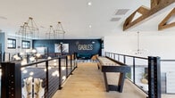The Gables is a thoughtfully designed amenity space that blends modern industrial aesthetics with inviting social areas across a multi-level layout. The upper level serves as the primary gathering space, finished with warm light hardwood flooring and defined by sleek black cable railings that overlook the dramatic lower level below. A bold navy accent wall bearing "The Gables" signage anchors a comfortable lounge area furnished with a teal velvet sofa, cognac leather chairs, and a chess table atop a layered area rug.

The game area is well-equipped, featuring both a shuffleboard table and a foosball table positioned along a charcoal accent wall adorned with sculptural palm leaf artwork. A high-top bistro table with industrial green metal stools offers additional casual seating near large windows that invite natural light throughout. Exposed wood beams add a warm contrast to the crisp white ceilings, while an array of gold hoop pendant lights and cascading globe pendants create a visually stunning focal point above the open stairwell. Dark textured tile cladding on a central column adds depth and architectural interest, completing the polished yet relaxed ambiance of this communal gathering space.