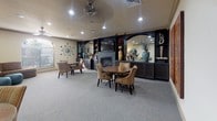 This single-floor community lounge is designed to provide a comfortable and versatile gathering space. The room is finished with neutral-toned walls and plush wall-to-wall carpeting, creating a warm and inviting atmosphere throughout. Recessed lighting and a ceiling fan illuminate the space evenly, complementing the overall layout.

The main lounge area is anchored by a striking dark built-in wall unit that houses a flat-screen television, a fireplace, open display shelving with decorative accents, and a mirrored backsplash. Several round dining tables, each surrounded by striped upholstered chairs, are arranged throughout the room, offering multiple intimate seating arrangements ideal for socializing or casual dining.

A cozy alcove off the main area features a plush sectional sofa, teal ottoman accents, a bold abstract canvas, and a statement floor lamp, providing a more relaxed lounge setting. Bold, colorful artwork adorns the walls throughout, adding visual interest and character to the space.

An adjacent kitchenette, visible through a connecting doorway, includes dark cabinetry, countertop appliances, and a sink, offering convenient service support for gatherings and events.