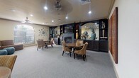 This single-floor community lounge is designed to provide a comfortable and versatile gathering space. The room is finished with neutral-toned walls and plush wall-to-wall carpeting, creating a warm and inviting atmosphere throughout. Recessed lighting and a ceiling fan illuminate the space evenly, complementing the overall layout.

The main lounge area is anchored by a striking dark built-in wall unit that houses a flat-screen television, a fireplace, open display shelving with decorative accents, and a mirrored backsplash. Several round dining tables, each surrounded by striped upholstered chairs, are arranged throughout the room, offering multiple intimate seating arrangements ideal for socializing or casual dining.

A cozy alcove off the main area features a plush sectional sofa, teal ottoman accents, a bold abstract canvas, and a statement floor lamp, providing a more relaxed lounge setting. Bold, colorful artwork adorns the walls throughout, adding visual interest and character to the space.

An adjacent kitchenette, visible through a connecting doorway, includes dark cabinetry, countertop appliances, and a sink, offering convenient service support for gatherings and events.