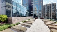 This sophisticated outdoor terrace is positioned at an elevated level within a dense urban environment, offering a striking juxtaposition of open-air relaxation amid a dramatic cityscape. The terrace is finished with large-format light gray porcelain pavers that lend a clean, contemporary aesthetic underfoot. A series of low-profile wooden daybed loungers with neutral cushions are arranged in organized rows, providing generous seating for relaxation and socializing. Raised wooden planter beds line the perimeter, filled with ornamental grasses and low-maintenance greenery that introduce natural texture and softness to the hard urban surroundings. Small circular accent tables punctuate the seating arrangements, adding practical functionality. A partial glass balustrade along one side maintains unobstructed sightlines to the surrounding skyline, while a textured wood-panel privacy wall provides shelter and visual contrast. The space is entirely open to the sky, maximizing natural light throughout the day. Surrounding glass curtain-wall towers reflect the city's skyline, creating a dynamic, ever-changing visual backdrop that defines the terrace's distinctive urban ambiance.