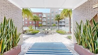 Located in Orlando's Dr. Phillips area, this outdoor amenity space at 5885 Lakehurst Dr offers a thoughtfully designed courtyard experience set within a multi-building residential community. The grounds are defined by a resort-style swimming pool enclosed by white fencing, surrounded by lounge chairs and mature tropical palm trees that establish a Florida-inspired ambiance throughout.

A covered pavilion anchors one end of the courtyard, featuring string lights overhead, a striped area rug, and ample seating — ideal for casual gatherings. Adjacent to the pavilion is a well-appointed outdoor kitchen with stainless steel grills set into a stone-faced counter, accompanied by a ping pong table, making it a versatile hub for recreation and entertaining.

Open green lawn areas provide generous open space, accented by decorative stone mulch beds and Florida-native plantings. Paved walkways connect the various zones of the courtyard, while white fencing neatly delineates the pool area and building perimeters. A covered outdoor seating nook with string lights and a dining table offers a more intimate setting. The multi-story buildings surrounding the courtyard feature private balconies overlooking the grounds, contributing to a sense of community and activated outdoor living throughout the space.