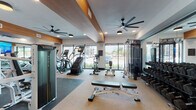 This fitness center is a thoughtfully designed, single-floor facility that offers a comprehensive workout environment for users of all fitness levels. The space is entered through glass double doors with sleek dark metal frames, setting a polished, professional tone from the moment of arrival.

Inside, the layout is divided into distinct functional zones. The cardio area lines the perimeter near floor-to-ceiling windows, housing a stair climber, elliptical trainers, upright and recumbent stationary bikes, and a row of Matrix treadmills. The expansive glazing floods the space with natural light and provides open views of the surrounding outdoor environment, creating an airy, energizing atmosphere.

The strength training section features a Matrix cable and weight machine, an adjustable bench, and a dedicated free weights zone with a full dumbbell rack. A functional training station offers medicine balls, kettlebells, and additional accessories neatly organized on a rack. Full-length mirrors line one wall, enhancing the sense of space and allowing users to monitor their form.

Overhead, large statement ceiling fans provide airflow throughout the space, complemented by recessed lighting. Warm wood-toned accent beams on the ceiling add an architectural element that contrasts with the otherwise contemporary, neutral palette of white walls and mixed flooring — carpet in the cardio zone and dark rubber flooring in the strength areas. The overall ambiance is modern, clean, and purpose-built for an effective fitness experience.