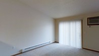 This single-floor space presents a clean, minimalist interior designed for functional living. The main room is characterized by its neutral color palette, with warm beige walls and a textured ceiling that complement the soft, plush wall-to-wall carpeting underfoot. The room offers a generous sense of openness, with smooth, uninterrupted wall surfaces that provide flexibility for furniture arrangement.

A prominent feature of the space is the large sliding glass door positioned along one wall, fitted with full-length vertical blinds that allow for adjustable light control. When the blinds are open, the door invites an abundance of natural light into the room, creating a bright and airy atmosphere. The sliding door also likely provides access to an outdoor area, adding a sense of connectivity to the exterior.

Climate comfort is addressed through a baseboard heating unit that runs along the base of the main wall, offering an efficient and unobtrusive heating solution. The overall ambiance of the space is calm and neutral, providing a versatile blank canvas well-suited to a variety of interior styles and personal preferences.