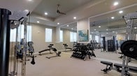 This fitness center is thoughtfully designed to support a comprehensive workout experience. The space features a tray ceiling with recessed lighting and a ceiling fan, ensuring a well-lit and comfortable environment during exercise. Large floor-to-ceiling mirrors line one wall, visually expanding the room and allowing users to monitor their form. Natural light filters in through multiple windows along the exterior wall, enhancing the bright, open atmosphere. The flooring is a neutral-toned carpet, providing a practical surface throughout the space. Equipment is well-organized and includes a dumbbell rack stocked with a range of free weights, adjustable workout benches positioned for versatile use, and a cable resistance machine visible toward the rear of the room. A framed piece of artwork adds a subtle decorative touch against the mirrored wall. The neutral beige wall tones and clean layout contribute to a functional yet welcoming ambiance, making this fitness center a practical and appealing amenity for users seeking a well-rounded exercise space.