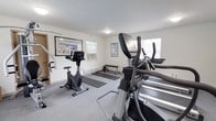 The fitness center at Woodbridge Terrace Apartments is a dedicated, single-floor facility designed to support a well-rounded exercise routine. The space is finished with neutral white walls, crown molding, and low-pile gray carpeting throughout, creating a clean and functional environment. Multiple windows allow natural light to fill the room, contributing to an open and comfortable atmosphere, while baseboard heating ensures year-round usability.

The cardio area is well-stocked with True-brand treadmills, an elliptical trainer, and an upright stationary bike, providing a range of options for aerobic workouts. A multi-station cable machine anchors the strength training section, offering versatility for resistance-based exercises. The fitness center also features a flat weight bench and yoga or exercise mats laid out on the floor, accommodating stretching, bodyweight training, and floor-based routines.

Instructional fitness posters and framed artwork decorate the walls, adding a polished, welcoming touch to the utilitarian space. Flush-mount ceiling light fixtures provide consistent overhead illumination throughout. Overall, the fitness center at Woodbridge Terrace Apartments presents a practical, comfortable, and comprehensively equipped amenity for residents seeking convenient on-site exercise options.