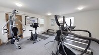 The fitness center at Woodbridge Terrace Apartments is a dedicated, single-floor facility designed to support a well-rounded exercise routine. The space is finished with neutral white walls, crown molding, and low-pile gray carpeting throughout, creating a clean and functional environment. Multiple windows allow natural light to fill the room, contributing to an open and comfortable atmosphere, while baseboard heating ensures year-round usability.

The cardio area is well-stocked with True-brand treadmills, an elliptical trainer, and an upright stationary bike, providing a range of options for aerobic workouts. A multi-station cable machine anchors the strength training section, offering versatility for resistance-based exercises. The fitness center also features a flat weight bench and yoga or exercise mats laid out on the floor, accommodating stretching, bodyweight training, and floor-based routines.

Instructional fitness posters and framed artwork decorate the walls, adding a polished, welcoming touch to the utilitarian space. Flush-mount ceiling light fixtures provide consistent overhead illumination throughout. Overall, the fitness center at Woodbridge Terrace Apartments presents a practical, comfortable, and comprehensively equipped amenity for residents seeking convenient on-site exercise options.