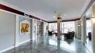 This single-floor lobby space exudes a classic, professional ambiance through its thoughtfully designed architectural details and refined finishes. The expansive entryway is anchored by gleaming stone tile flooring in cool grey tones, which reflects the overhead recessed lighting and contributes to the open, airy feel of the space.

A built-in reception desk is prominently positioned along one wall, framed by a decorative peaked archway and accented with warm wall sconces. This staffed station serves as the central point of contact for visitors and residents alike. Adjacent to the reception area, a set of glass-paneled doors provides a clearly marked interior passage, while potted greenery adds a touch of natural warmth.

Structural columns with detailed capitals punctuate the space, complementing the crown molding and the deep burgundy accent band that runs along the upper walls — a design choice that lends the lobby a distinguished, formal character.

Toward the front of the lobby, floor-to-ceiling windows and arched glazing flood the space with natural light while offering unobstructed views of the surrounding exterior. A seating area furnished with dark leather chairs and sofas provides a comfortable waiting area, making the lobby as functional as it is visually impressive.