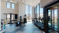 This contemporary fitness center is a standout amenity space defined by its dramatic architectural design and thoughtfully curated layout. The most striking feature is the expansive floor-to-ceiling glass curtain wall that floods the space with natural light and frames panoramic views of the surrounding urban skyline, including a prominent sports stadium and city landmarks visible in the distance.

The soaring double-height ceilings create an open, airy atmosphere enhanced by recessed lighting, ceiling-mounted fans, and clerestory windows that introduce additional light from above. A bold hexagonal tile floor pattern in tones of blue, grey, and taupe runs throughout the space, adding a modern, graphic element to the design.

The cardio zone is generously appointed with multiple treadmills, elliptical trainers, and a stationary bike, all positioned to take full advantage of the views. A dedicated strength and functional training area features a multi-station cable machine, a dumbbell rack, medicine balls, and a flat-screen TV mounted against a visually arresting accent wall adorned with bold, pop-art-style graphic wallcovering. A compact refreshment station with a mini-refrigerator and built-in cabinetry completes the space, ensuring a fully self-contained fitness experience.