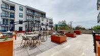 This outdoor courtyard terrace is nestled within the interior-facing side of a contemporary multi-story building, creating a semi-enclosed open-air environment that is both functional and visually striking. The terrace is finished with large-format concrete pavers that provide a clean, durable surface across the expansive patio area.

A defining design feature of the space is the series of oversized, bold orange steel planter boxes positioned throughout the terrace. These raised planters contain ornamental grasses and drought-tolerant plants, adding texture and vibrant contrast against the neutral tones of the surrounding architecture. A pergola-style steel framework overhead in portions of the courtyard adds architectural interest and subtle shade definition without enclosing the open sky above.

The terrace is furnished with multiple round bistro tables and wire-frame chairs, providing ample seating for outdoor gathering and relaxation. A large market umbrella adds additional shade in one section of the patio. Black powder-coated metal railings delineate pathways and transitional zones within the terrace, lending a cohesive and modern aesthetic throughout.

The building's white stucco exterior, black-framed windows, and steel balconies frame the courtyard on multiple sides, reinforcing the contemporary design language. Open sky and distant mountain views are visible beyond the perimeter, enhancing the airy ambiance of this well-appointed outdoor space.