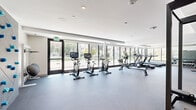 Located at 8377 Blackburn Ave in Los Angeles, CA, this well-appointed fitness center offers a comprehensive exercise facility designed to support a wide range of workout routines. The space is organized into distinct functional zones, making it easy to transition between cardio and strength training. Floor-to-ceiling windows line one side of the facility, flooding the interior with natural light and providing refreshing views of the surrounding greenery — creating an energizing atmosphere for workouts. The cardio zone features a row of stationary bikes equipped with digital screens, treadmills, a rowing machine, and an elliptical trainer, all positioned to take advantage of the outdoor views. The strength training area includes a power rack with an extensive weight plate storage system, a full dumbbell rack with a flat bench, wall-mounted mirrors to aid form, TRX suspension trainers, kettlebells, medicine balls, and a multi-functional cable machine. The flooring throughout consists of durable rubber gym flooring — lighter in the cardio zone and darker speckled rubber in the strength area — ensuring safety and comfort. Recessed lighting and a clean white palette give the space a modern, professional aesthetic, while restroom facilities are conveniently accessible on-site.