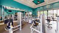 Located at 4600 Pear Grove Ct in Glen Allen, Virginia, this single-level fitness center is designed to support a comprehensive workout experience. The space is immediately distinguished by its vibrant teal walls, which are complemented by crisp white trim and elegantly coffered ceilings with recessed lighting and ceiling fans—lending the room a polished, upscale character.

The fitness floor is outfitted with a wide array of modern equipment, including Precor cardio machines such as ellipticals and treadmills, as well as a full selection of strength-training stations and cable machines. A dedicated free weights area features a dumbbell rack alongside an adjustable bench, accommodating a range of training preferences. Full-length mirrors line the walls, visually expanding the space and allowing exercisers to monitor their form throughout their sessions. Wall-mounted flat-screen televisions provide entertainment during workouts.

Natural light flows generously through large windows and expansive sliding glass doors, which open directly to an outdoor patio area featuring stone columns and views of a pool. Durable rubber flooring throughout ensures both safety and comfort. An adjacent room, visible through an interior doorway, suggests additional amenity space nearby. The overall ambiance is energetic yet refined, making this fitness center a functional and attractive facility.