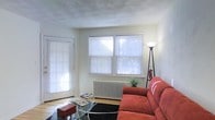 This single-floor space features a welcoming living area defined by its light-filled, airy atmosphere. Large windows dressed with horizontal blinds line the walls, allowing generous amounts of natural light to pour into the room while offering views of the surrounding greenery outside. A classic cast-iron radiator beneath the windows serves as a functional and period-appropriate heating feature, adding a touch of vintage character to the space.

The living area is finished with hardwood flooring, partially anchored by a dark area rug that grounds the seating arrangement. A bold red sofa provides a vibrant focal point, complemented by a glass-top coffee table that keeps the look light and open. A tripod floor lamp adds both style and supplementary lighting to the corner of the room.

The ceiling features a subtle textured finish, adding visual interest overhead. A door with an accompanying window along one wall provides an additional source of natural light and suggests access to an exterior area. The overall palette of the space is neutral and clean, with white walls creating a versatile backdrop that enhances the sense of openness throughout.