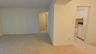 This single-floor residential space presents a straightforward and functional layout designed for comfortable everyday living. The interiors are finished in a clean, neutral color palette, with white walls and light-toned carpeting throughout, creating a bright and cohesive atmosphere.

The main living area is open and generously proportioned, offering flexible arrangement possibilities. A partial wall adds subtle definition to the space without compromising the open feel. The room flows naturally into a hallway that leads deeper into the unit, maintaining a sense of connectivity between areas.

At the end of the hallway, a built-in shelving unit provides practical storage space, ideal for books, décor, or organizational needs. This feature adds a functional architectural element to an otherwise clean and unadorned interior.

The space benefits from a simple, uncluttered design throughout, with smooth ceilings and consistent flooring that tie the rooms together seamlessly. The neutral finishes offer a versatile backdrop that can accommodate a wide range of furnishing styles and personal touches, making it an adaptable and move-in ready living environment.